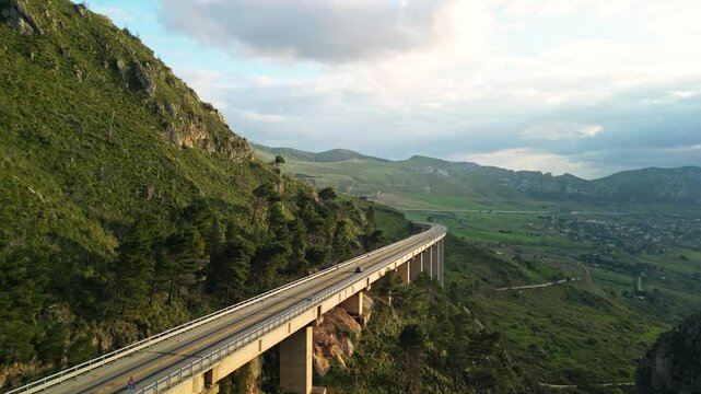 Road bridge spanning a mountainous landscape at sunset, showcasing vehicles traveling along the elevated structure with lush greenery and hills in the background