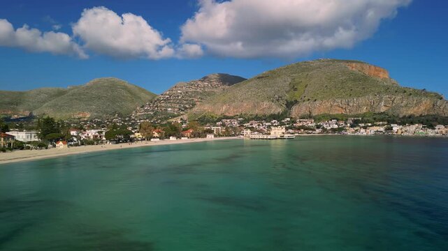 Mondello Beach. Scenic view of Mondello Beach with clear turquoise water, sandy shore, and lush green hills under a bright blue sky with fluffy clouds