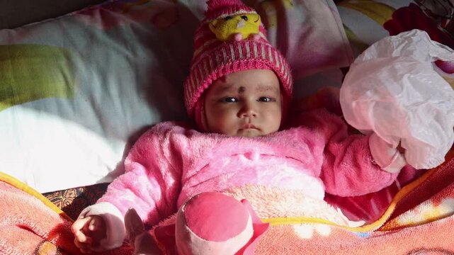 A baby girl lies on a bed wearing warm winter clothes, looking calmly at the surroundings, capturing a peaceful and tender childhood moment in a simple rural village setting