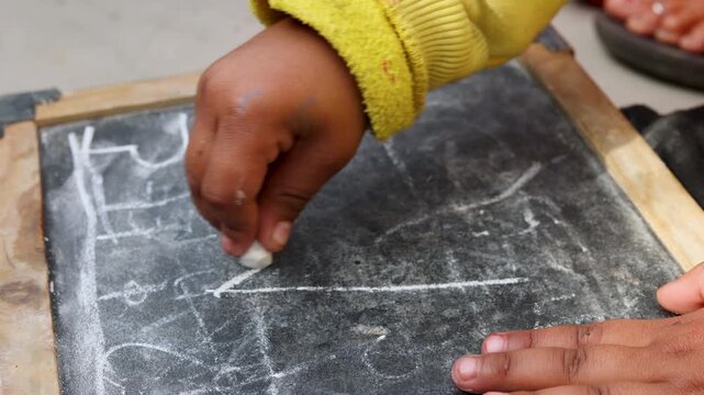A young Indian child uses chalk to draw on a slate board, capturing a simple learning and creative moment of childhood education in a rural village setting