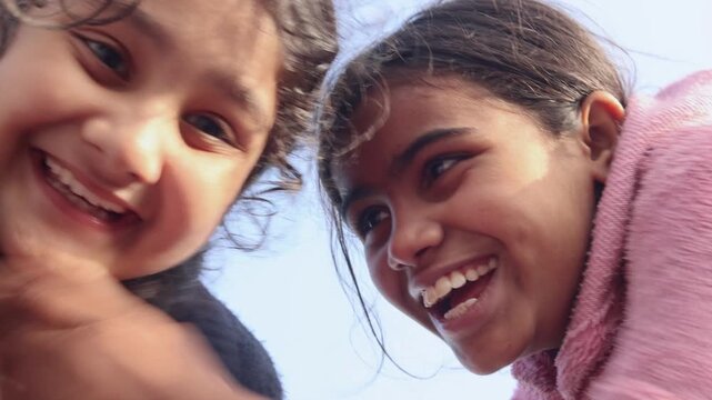 Two young Indian girls smile brightly while looking down toward the camera, capturing a joyful and playful childhood moment with natural expressions in a simple rural village setting