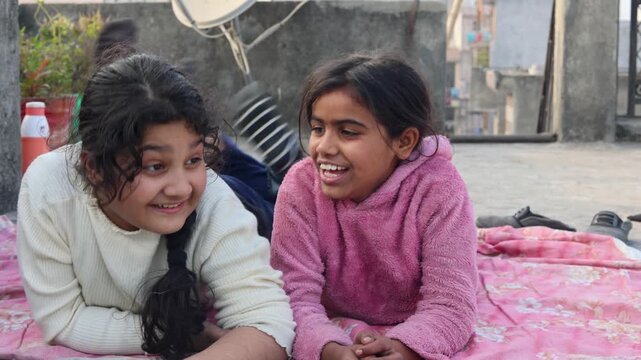 Two cheerful Indian village girls lying together and smiling during a playful conversation, capturing friendship, happiness, childhood innocence, and rural lifestyle in Uttarakhand mountain setting.