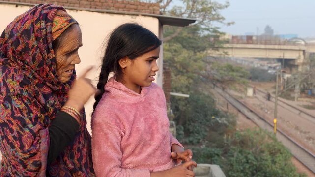 An elderly woman and a young girl standing together outdoors, looking into the distance, capturing a quiet generational bond and thoughtful moment in a modest urban neighborhood setting.