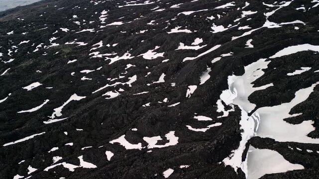 Aerial view of volcanic landscape with black lava rock and patches of white snow, showcasing the rugged terrain and natural formations in a remote area