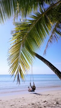 Experience the beauty of Koh Kood, Thailand, as a person enjoys a peaceful moment swinging beneath a palm tree. The stunning beach and clear blue waters create a perfect vacation backdrop.