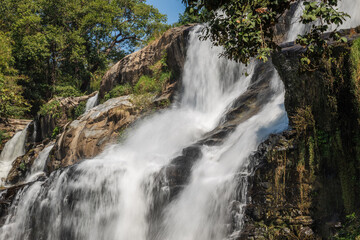 Obraz premium Experience the majestic cascade of Mae Ya Waterfall in Chiang Mai, Thailand, where rushing waters gracefully flow over multiple rock tiers, framed by vibrant tropical foliage under a clear sunny day.