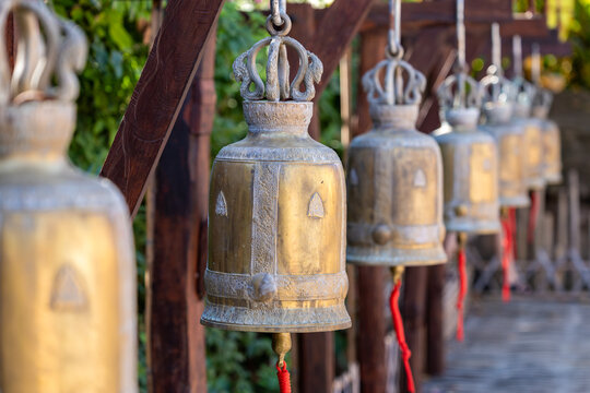 A row of ornate, aged brass temple bells with intricate carvings and red cords, hanging from wooden beams at Wat Saket in Bangkok, Thailand, evoking spiritual tranquility.
