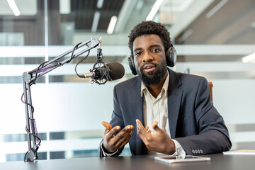 Naklejka premium Young man broadcasting live, creating content for podcast or radio show, talking and gesturing with hands while wearing a suit and headphones in a modern studio