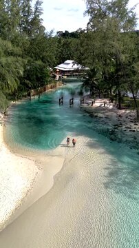 Two people stroll along the soft sandy shore of a tranquil lagoon in Koh Kood Island, Thailand. Lush greenery surrounds the serene blue waters, creating a peaceful tropical paradise.
