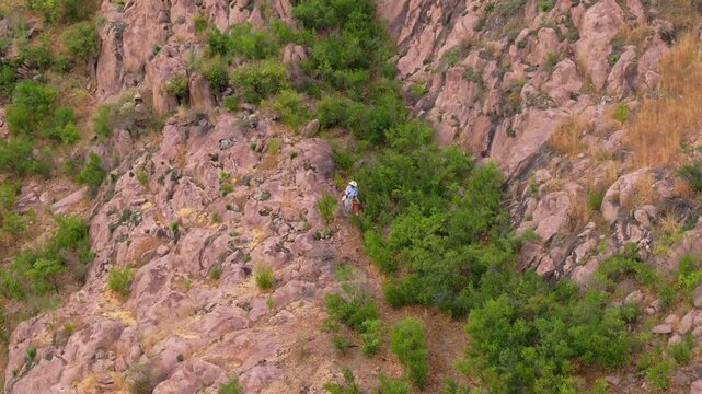 Aerial footage of man on horse navigating through difficult rocky cliff edge at height. Pink and brown boulders contrast with vibrant green desert shrubs dotting the rugged mountainside terrain.