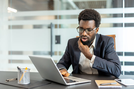 Businessman in spectacles looking at his laptop screen with a surprised and concerned expression, experiencing frustration and shock while working in a modern office environment