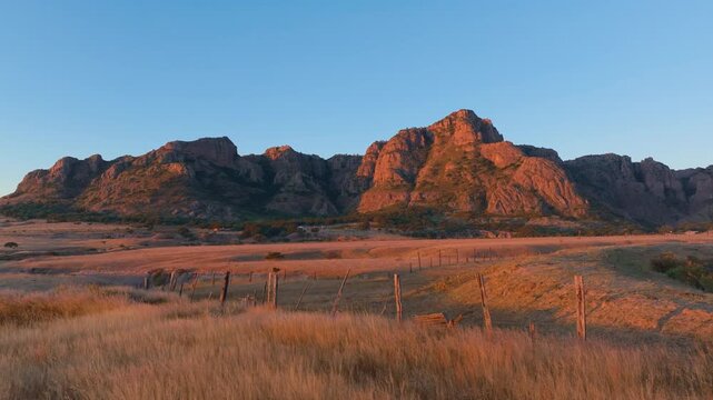 Majestic red rock mountains bathed in warm sunset light rise above golden dry grassland with rustic wooden fence posts in Durango, Mexico. Clear blue sky frames the rugged peaks and arid terrain.
