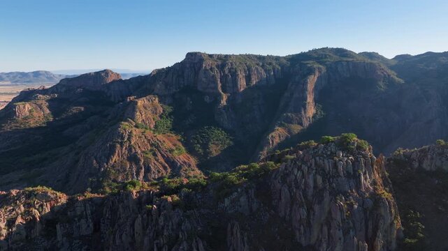 Drone footage reveals dramatic mountain landscape in Durango, Mexico featuring steep rocky cliffs, deep canyons, and scattered green vegetation under clear blue sky during golden hour light.