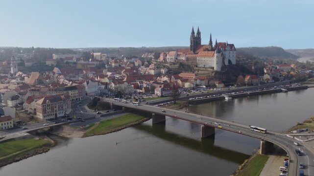 Wide-angle panorama of Meissen featuring the Elbe river and bridge. The Meissen Cathedral and Albrechtsburg Castle tower majestically over the historic old town under a clear blue sky.