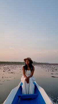 A tranquil moment unfolds as a woman in a white dress enjoys the stunning sunset at Lake Nong Harn, surrounded by vibrant red lotus flowers. Red Lotus Sea, Udon Thani, Thailand