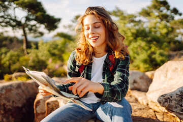 A hiker with a backpack stands on a mountain with a map in her hands. A young woman with a map looks for directions while hiking. Concepts of enjoyment, navigation. Active lifestyle. © maxbelchenko