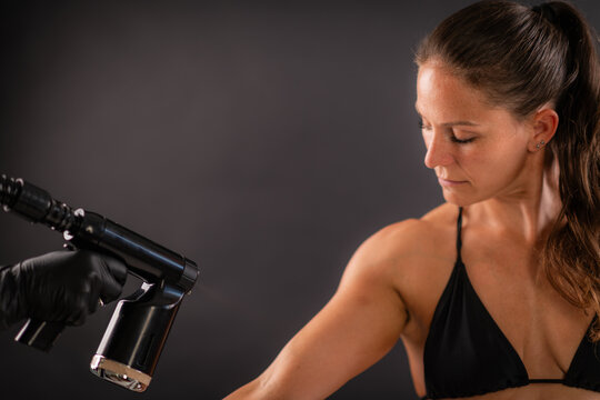 Applying a fine tanning mist to the upper arm using a professional spray device. Controlled technique supports even coverage and uniform cosmetic skin tone.