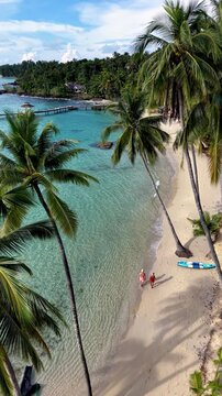 Explore the beautiful shores of Koh Kood Island in Thailand, where golden sands meet turquoise waters. Palm trees sway gently as two people stroll along the inviting beach under a sunny sky.