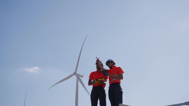 Asian engineers using a tablet and clipboard to inspect wind turbines at a renewable energy site, representing clean energy operations and technical teamwork in sustainable engineering.