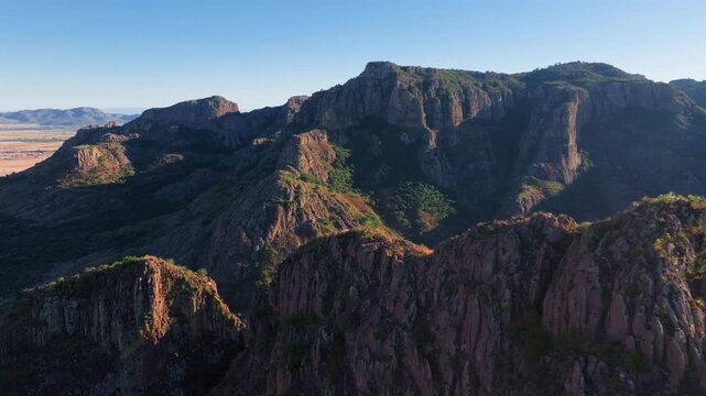 Breathtaking aerial drone footage reveals dramatic mountain landscape in Durango, Mexico with towering rocky cliffs, deep valleys, lush green vegetation patches, and warm golden morning sunlight.