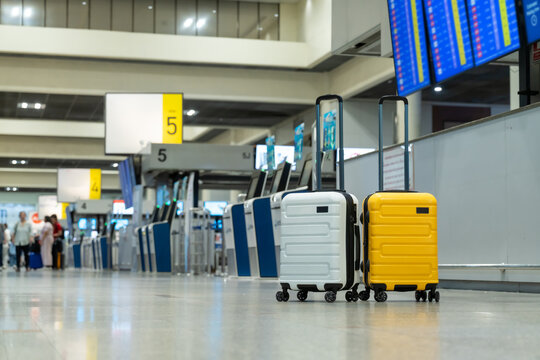 Two modern hard-shell suitcases, yellow and white, are standing in a blurred airport terminal departure hall near check-in counters and flight information displays, ready for travel and vacation.