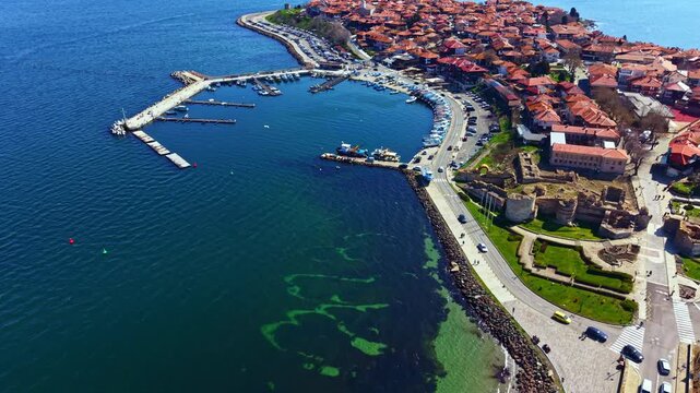 Visitors stroll through the streets of Nesebar Old Town, observing historic buildings and the marina filled with boats during a bright sunny day by the sea.