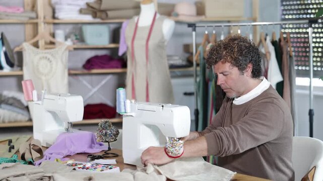 Adult man sews clothes on a sewing machine against the background of shelves with fabrics and a mannequin. Process of sewing a collection of clothes in the workshop