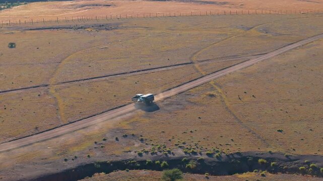 White pickup truck travels along dusty dirt road through expansive rural farmland with golden dry grass fields and wooden fence posts. Mountains visible on distant horizon under clear sky.