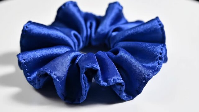Vibrant blue ruffled satin fabric scrunchie hair accessory centered on a plain white surface with soft shadows, showcasing the shiny textile texture and elastic circular design.