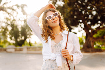 A young woman stands by an old stone wall in the city. She wears sunglasses and a light outfit while holding a handbag. The sun shines bright and people walk in the background. © maxbelchenko