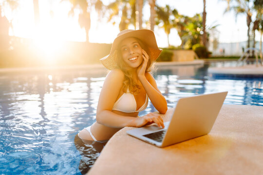 Happy woman stands in pool, working on laptop from parapet on a sunny day. A female freelancer relaxes by the pool with a laptop at sunset. Concept of remote work, relaxation, and enjoyment.