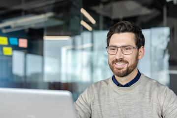 Obraz premium Happy professional man wearing casual sweater and eyeglasses smiling while using a laptop, representing success, productivity, and positive working environment