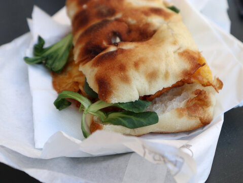 Panino gourmet con focaccia croccante farcita con ingredienti freschi e valeriana, servito in carta alimentare bianca, concetto di street food italiano e pranzo veloce