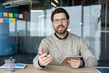 Naklejka premium Smiling business professional using a notepad while participating in an online meeting, looking at the camera and explaining a business concept during a remote work conference