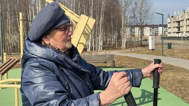 Woman exercising outdoors spring stadium wearing glasses beanie coat using leg exerciser on green rubber ground steady alternating.