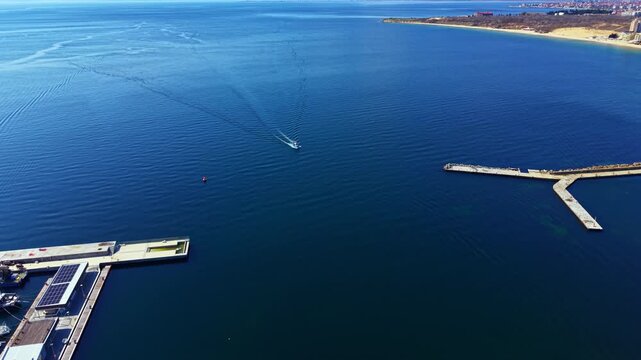 A boat travels over calm water close to a marina. The shore is visible in the distance, with land and structures outlined against the blue sky during daylight.