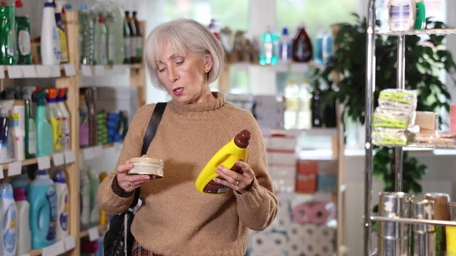 Attentive old woman reviewing parquet and wood cleansing agents in household goods shop 