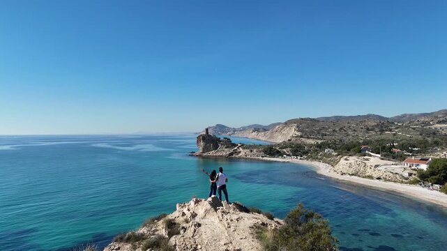 Cinematic low drone flight over couple watching ruined coastal tower above cliff and sea