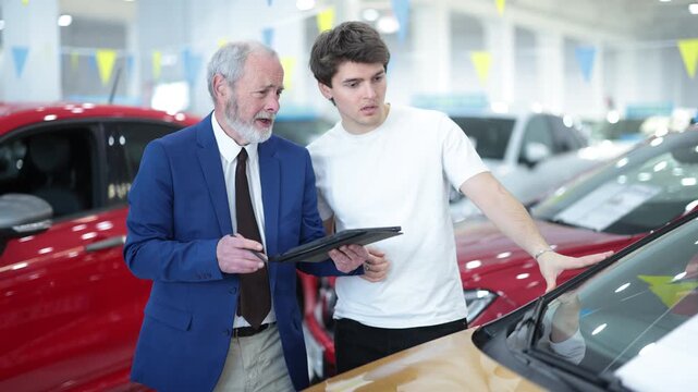 Elderly man seller shows the car to a guy who is a client of the car dealership. Experienced consultant sells a comfortable automobile on favorable terms. High quality 4k footage