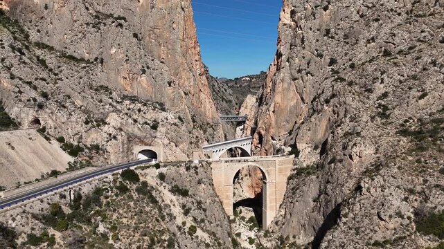 Ascending orbital drone shot around Mascarat Canyon bridges with road traffic