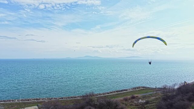 A paraglider glides above the calm water near the coast. The person enjoys the view of the sea and distant hills. The sky shows some clouds and the bright sunlight makes the scene lively.