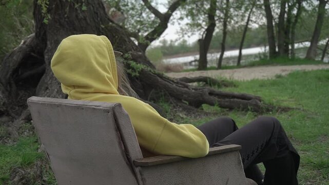a girl in a yellow hoodie sits motionless in an old vintage chair, hiding her hands in her pockets and admiring the beauty of the local nature outside the city, against the backdrop of a large thick