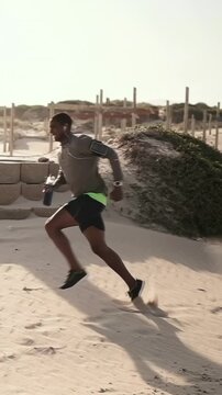 Vertical video: Tracking runner running across beach kicking sand with bottle training in grey top