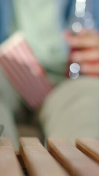 Vertical video: Seeing remote, denim man nudging remote on slat table to control TV holding bottle