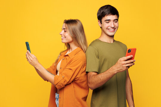 Side view young couple two friends family man woman wear casual clothes hold in hand use mobile cell phone together stand back to back isolated on pastel plain light yellow background studio portrait.