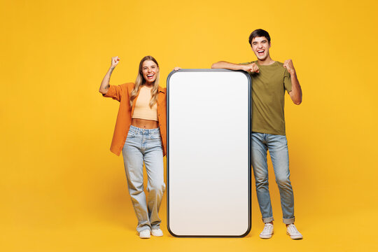 Full body young couple friends family man woman wear casual clothes together big huge blank screen mobile cell phone smartphone with area do winner gesture isolated on pastel plain yellow background.