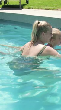 Vertical video: Pointing mom in olive suit guiding girl using blue kickboard at pool, gaining skill