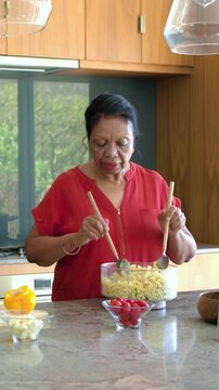 Vertical video: Senior woman grasping spoons, stirring bowl of salad as friend chopping, cooking