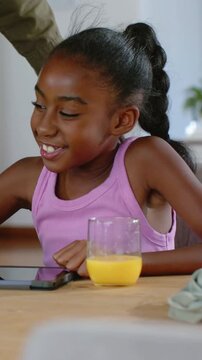 Vertical video: Girl using stylus on tablet at table for learning, adult guiding, orange juice