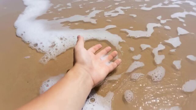Extending bare forearm and open hand over wet sand while incoming wave curling foam around fingers